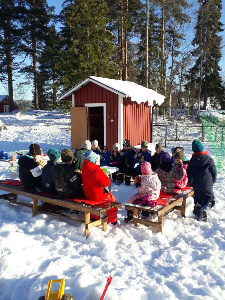 Barn sitter i ring runt eldstad i snön.